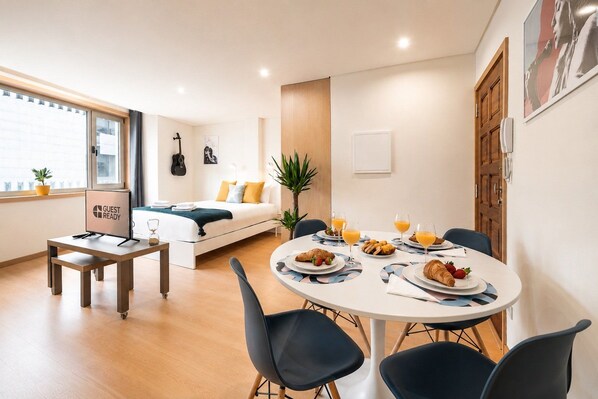 A modern hotel room with a bed, a dining table with breakfast, a guitar, and a 'Guest Ready' sign.
