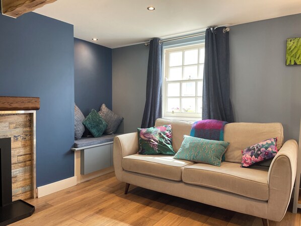 A cozy living room with a beige sofa, a wooden fireplace, and a window with curtains. - The Ship Inn Self Catering (Castle Douglas)