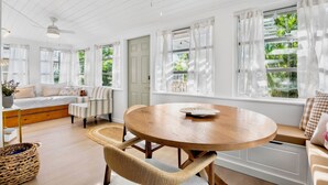 A bright, airy room with a wooden floor, a round wooden table, a bed with a bench, and large windows with sheer curtains. - Tiny Tides: Adorable Cozy House! (Anna Maria)