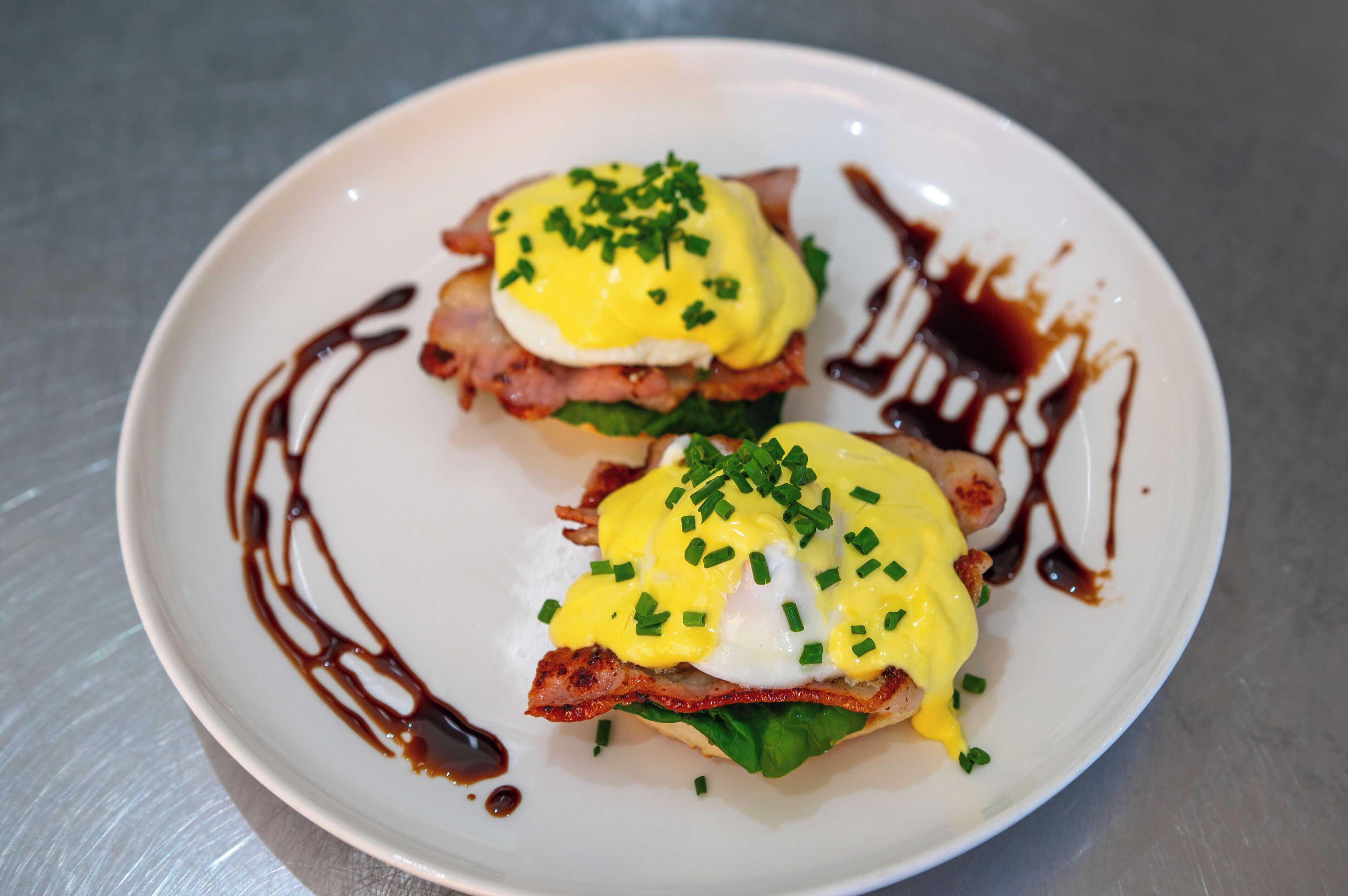Un plato con dos sándwiches eggs benedict cubiertos de tocino, salsa holandesa y cebollín.