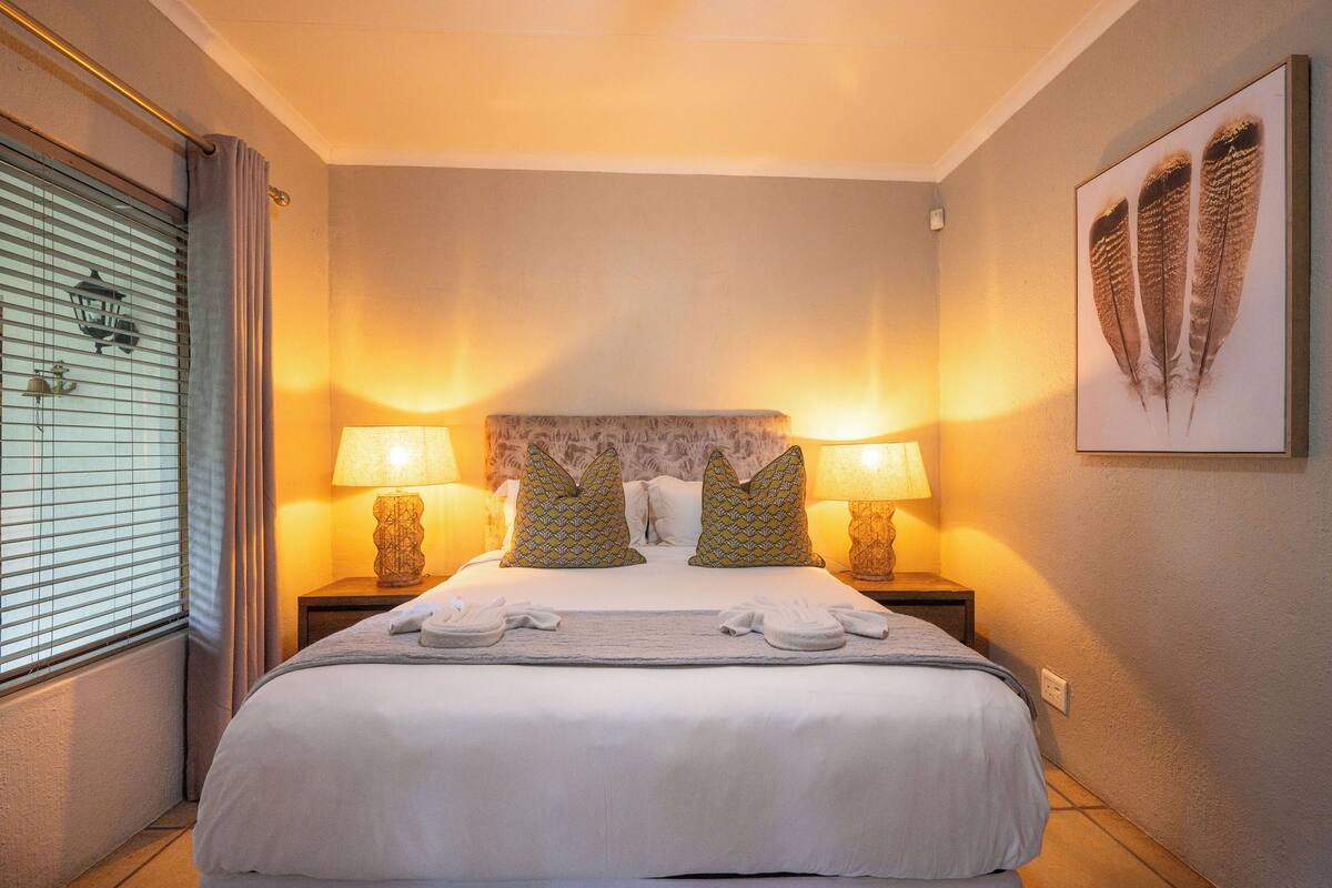 Ein Hotelzimmer mit einem großen Bett, zwei Nachttischlampen, einem Kopfteil mit einem dekorativen Muster, einem Wandbild und einem Fenster mit Jalousien.