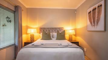 A hotel room with a large bed, two bedside lamps, a headboard with a decorative pattern, a wall-mounted artwork, and a window with blinds.