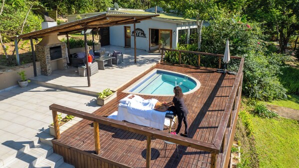 A pool area with a wooden deck, a lounge chair, and a person standing by the pool.