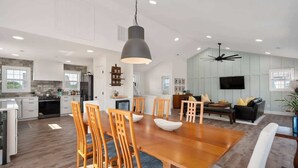 Una cocina amplia con armarios blancos, un comedor con mesa y sillas de madera, y una sala de estar con sofá y televisor.