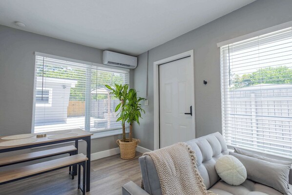 A living room with a grey sofa, a small table, a potted plant, and large windows with blinds.
