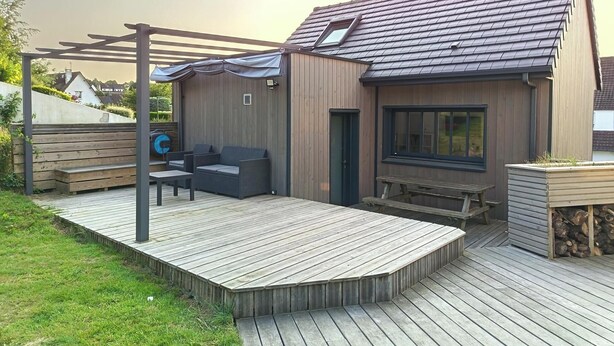A modern outdoor deck with a built-in seating area, a pergola, and a small building with a sloped roof. - Wooden cottage (Longuenesse)