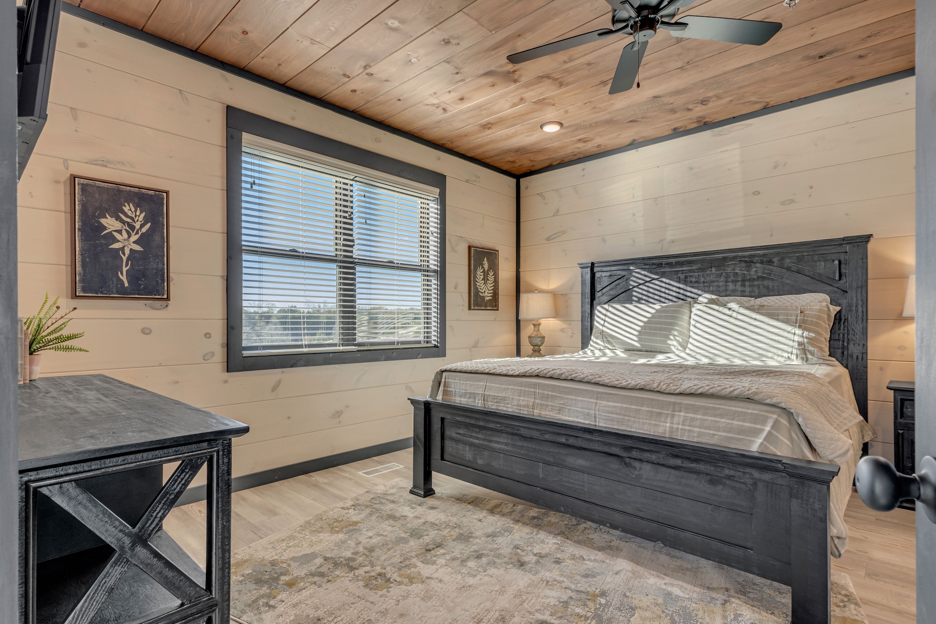 A bedroom with a wooden ceiling, a bed with a dark headboard, and a window with blinds.