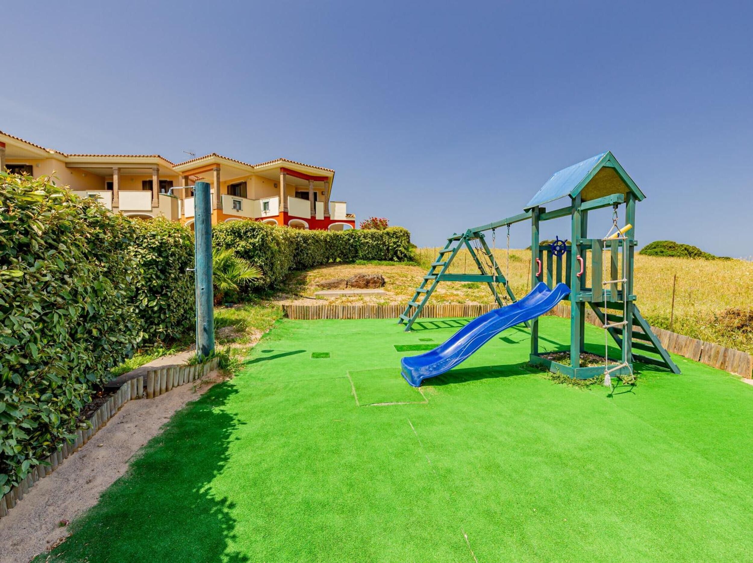 A children's play area with a slide and climbing structure, surrounded by well-maintained grass and a hedge.