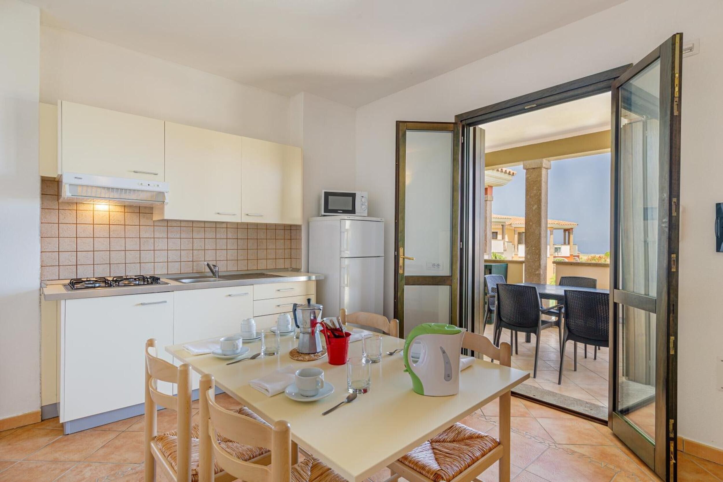 A compact kitchen with white cabinets, a gas stove, and a microwave. There is a dining area with a table set for two, and a sliding glass door leading to an outdoor seating area.
