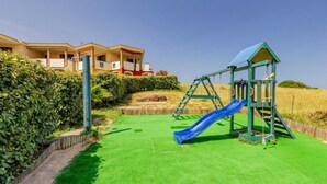 A playground with a blue slide and a climbing structure, surrounded by green artificial grass, with a building in the background.