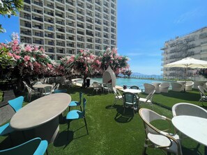 Uma área de pátio externa com mesas, cadeiras, piscina e vista para o mar.