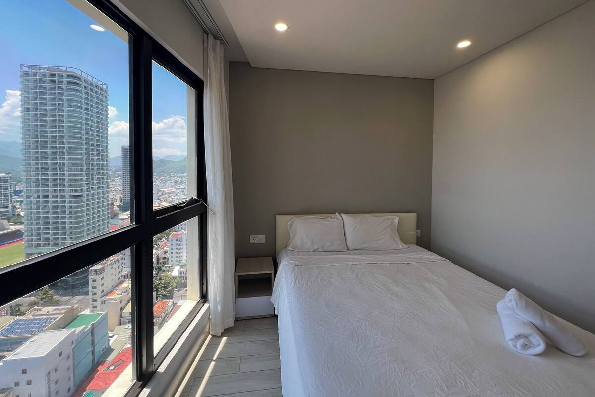 Una habitación de hotel con un ventanal grande, una cama con ropa blanca y vistas a un paisaje urbano.