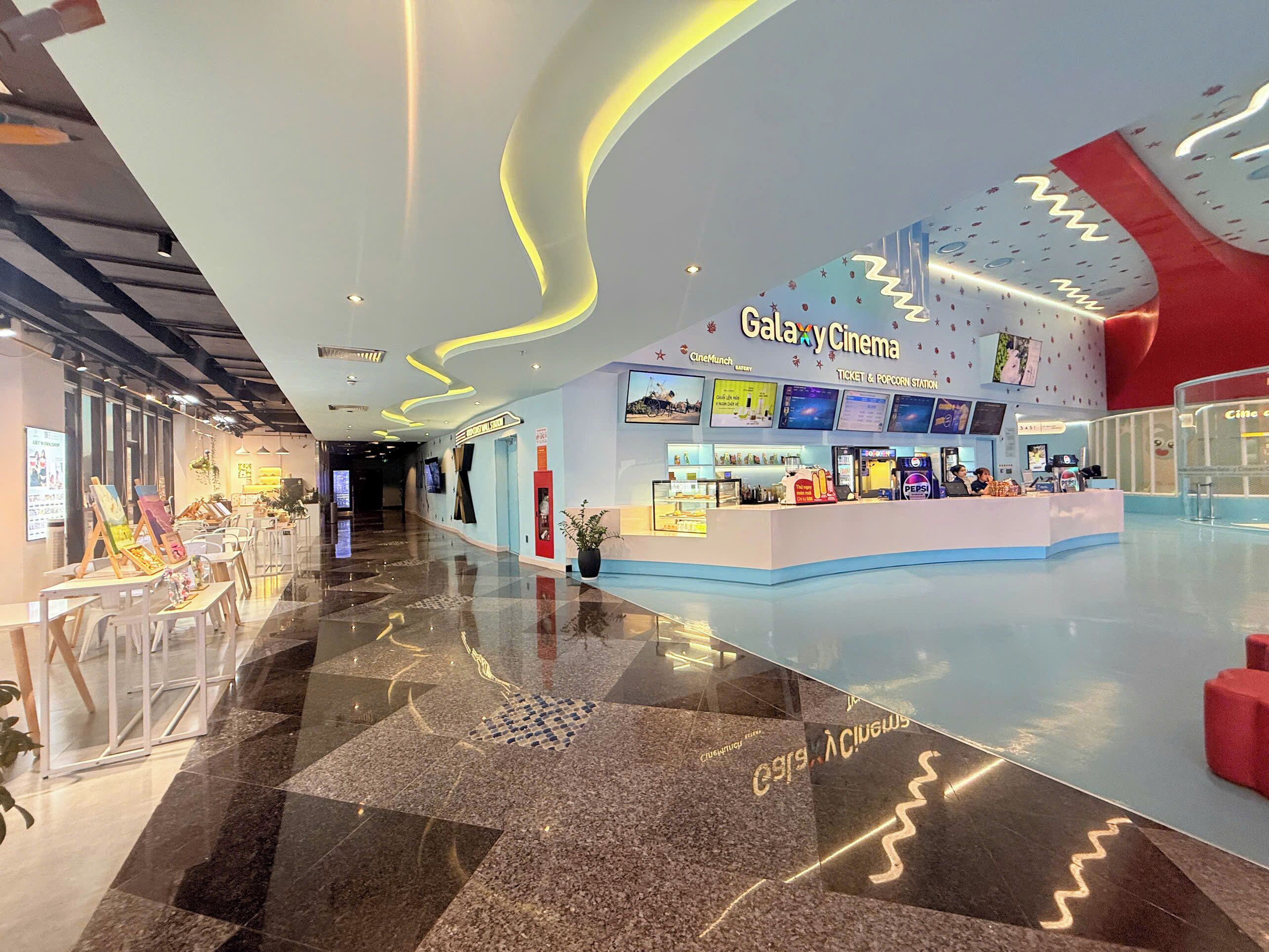A modern cinema with a ticket and popcorn station, a seating area with tables and chairs, and a decorative ceiling.