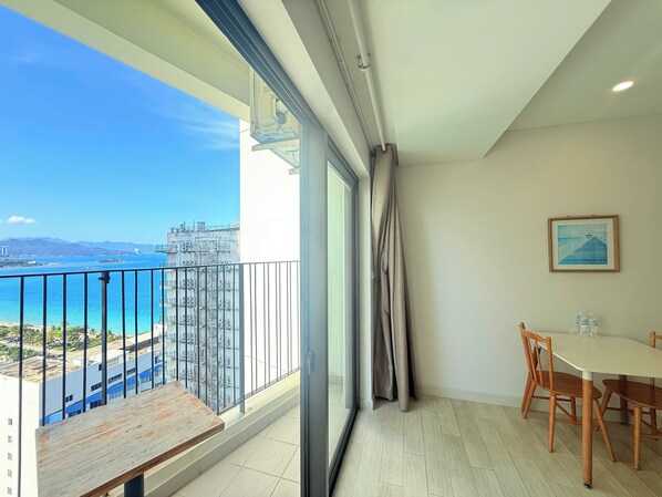 Un balcón con vista al mar y edificios, una mesa de comedor con sillas y un cuadro en la pared.