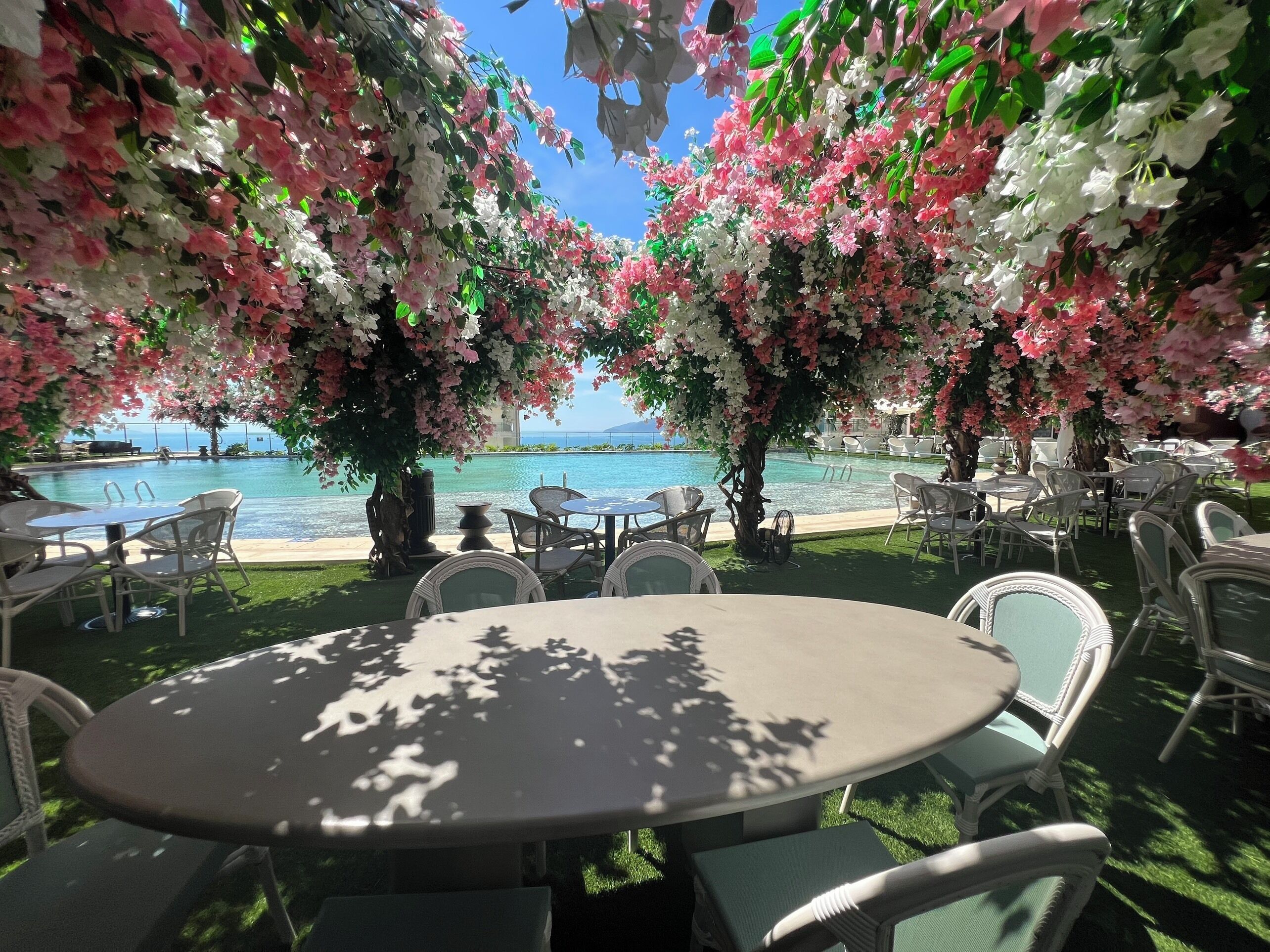An outdoor seating area with round tables and chairs, surrounded by lush pink and white flowering trees, overlooking a pool and a clear blue sky.