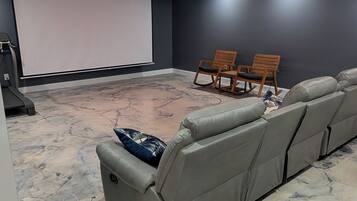 A home theater room with a large gray sectional sofa, a small table, two wooden chairs, and a large screen.