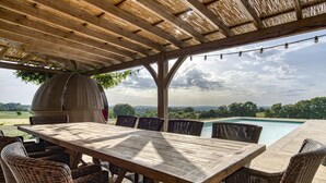 An outdoor patio with a wooden table and wicker chairs, overlooking a swimming pool and a scenic landscape.