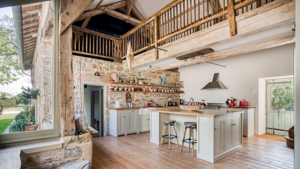 A rustic kitchen with a stone wall, wooden beams, and a large island with stools.