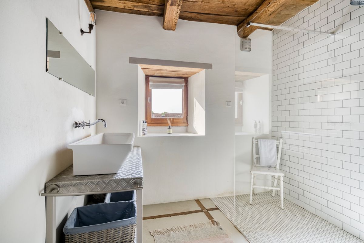A bathroom with a large white sink, a mirror, a window, and a white tiled shower area.