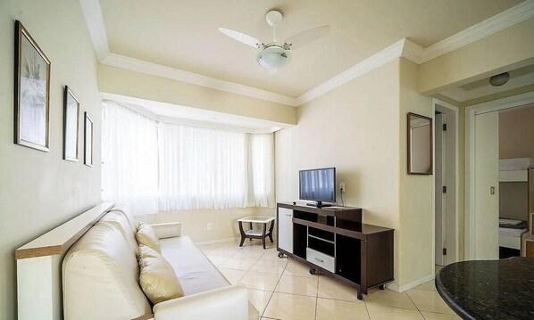Uma sala de estar com sofá, televisão sobre um móvel, ventilador de teto e janela com cortinas.