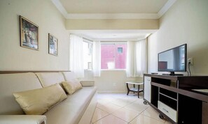 Sala de estar com sofá, televisão, mesa pequena e janela com vista para um edifício rosa.