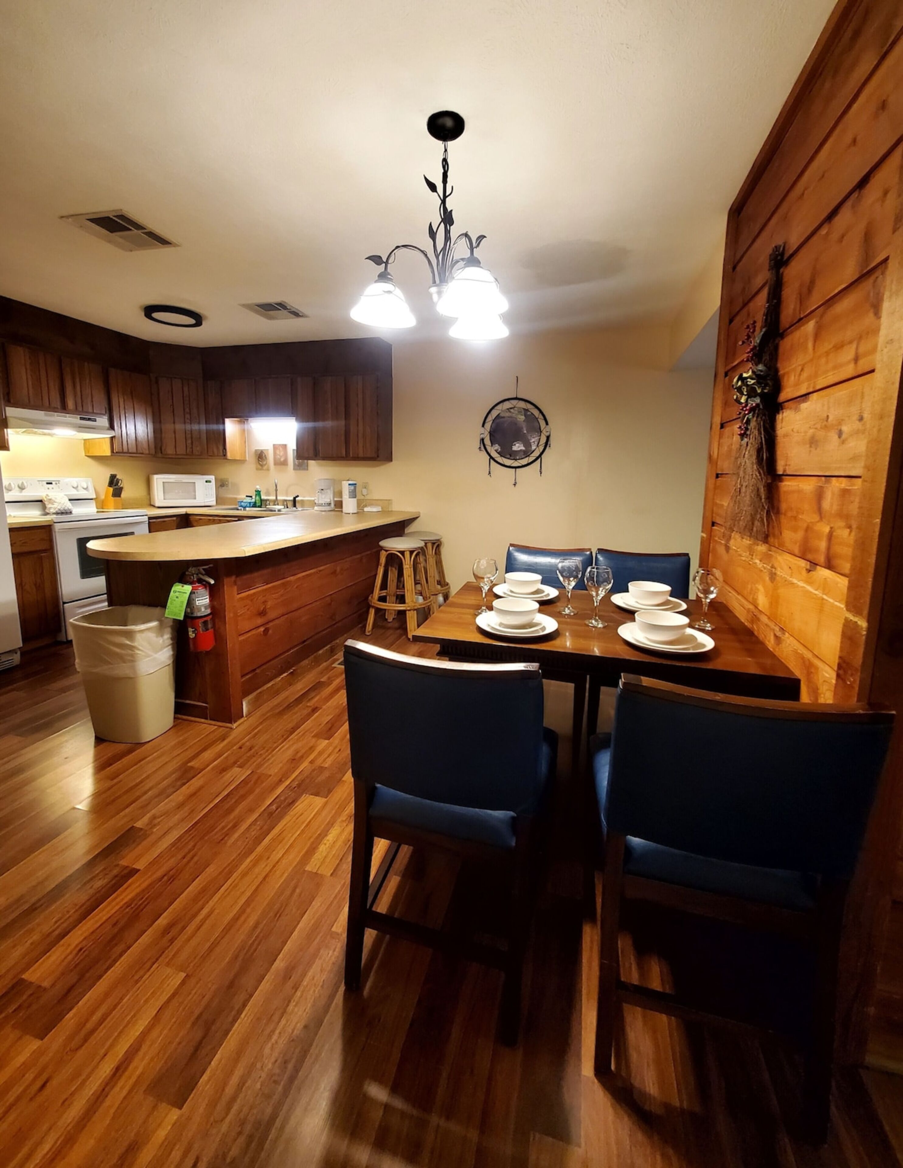 Une table de salle à manger en bois avec des chaises, un coin cuisine avec des meubles en bois, un micro-ondes blanc et un luminaire au plafond.