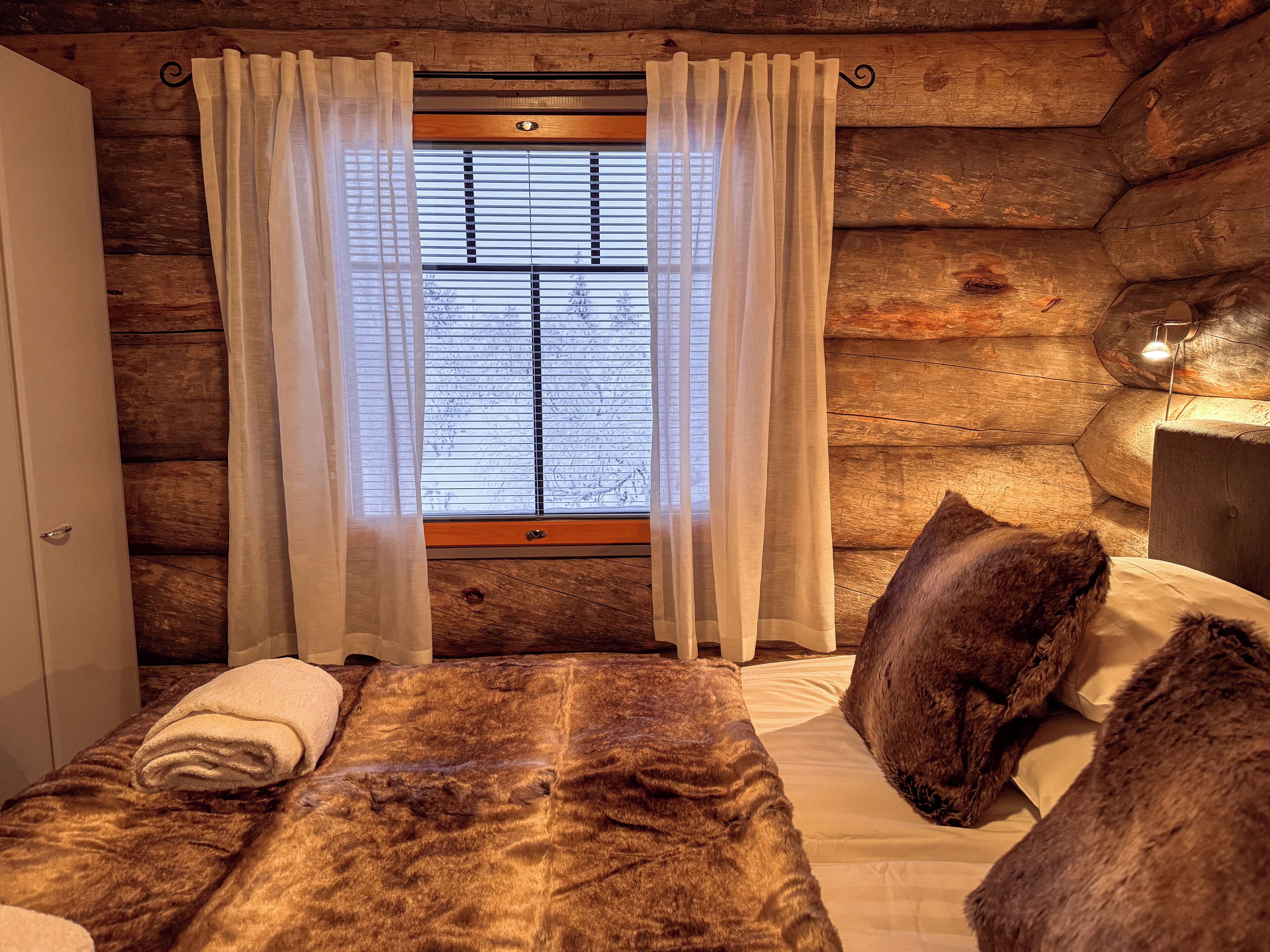 Una camera in una baita di legno con un letto, una finestra con tende leggere e un comodino con una lampada.