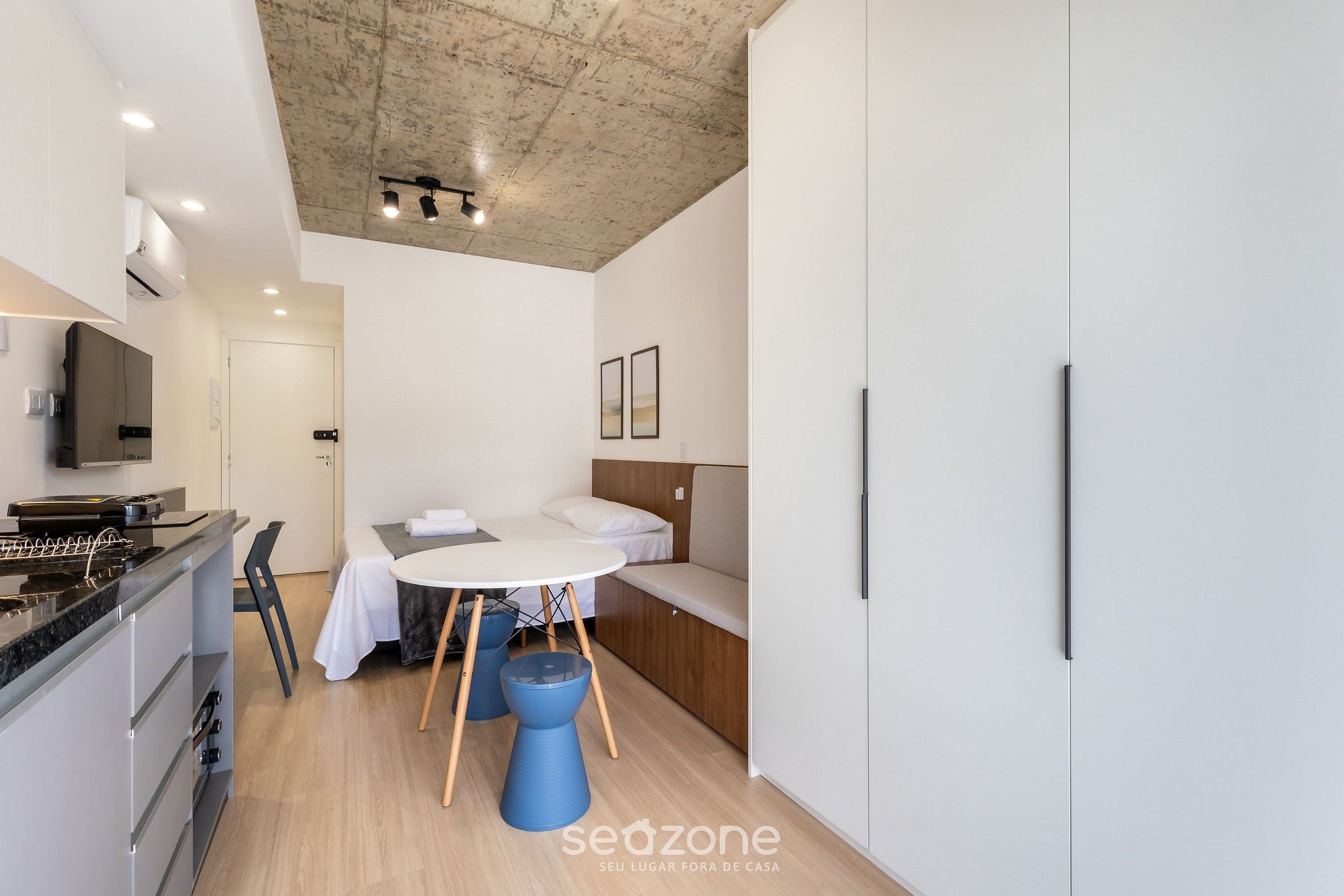 Quarto moderno e compacto com cozinha compacta, cama com encosto, mesa de jantar redonda e poltrona azul.