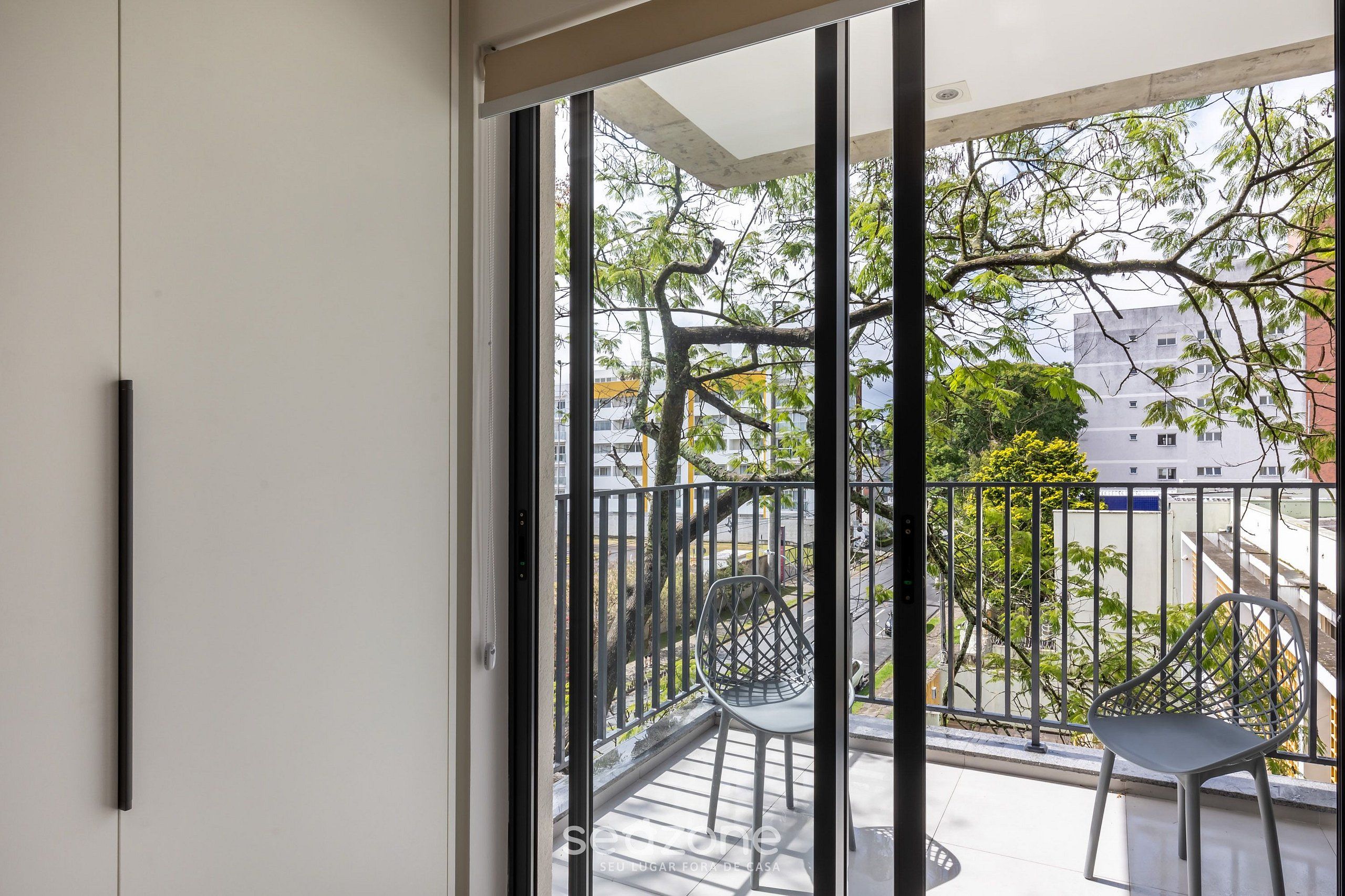 Un balcón con barandilla de metal, una silla y vistas a un árbol y edificios.