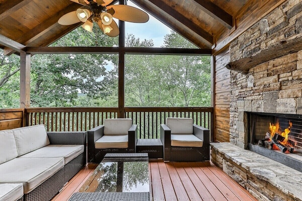 A covered deck with wicker furniture, a stone fireplace, and a view of trees.