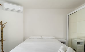 A neatly made bed with white linens and two rolled towels, a bedside table, a wall-mounted air conditioner, and a wooden decorative piece.