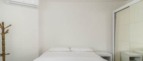 Una cama bien hecha con sábanas blancas y dos toallas enrolladas, una mesita de noche, un aire acondicionado de pared y un adorno de madera.