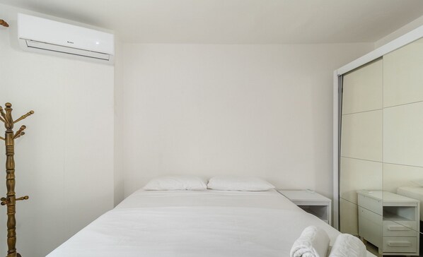 Una cama bien hecha con sábanas blancas y dos toallas enrolladas, una mesita de noche, un aire acondicionado de pared y un adorno de madera.