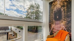 A room with a large window overlooking a road and trees, a chair with an orange throw, and a poster of a railway track. - Cozy Themed Apartment in Canela Dal221 (Canela)