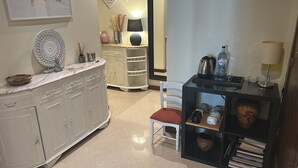 Una cocina con encimera de mármol, armarios blancos, una silla blanca y una mesita auxiliar negra con una botella de agua y una tetera.