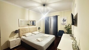 Habitación de hotel con una cama grande, un escritorio, una silla, un radiador, un espejo, una planta y un televisor.