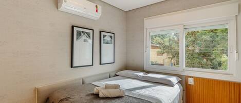 A bedroom with a bed, air conditioning unit, framed pictures, and a window with a view.