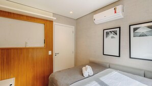 Quarto de hotel com porta de madeira, cama com cabeceira cinza, ar condicionado e duas imagens emolduradas na parede.
