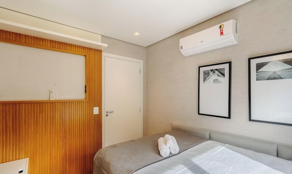 Quarto de hotel com cama, guarda-roupa de madeira, ar condicionado e duas imagens emolduradas na parede.