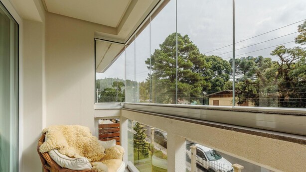 Uma varanda com cadeira de vime e almofada fofa, com vista para uma área residencial com árvores e um carro estacionado.