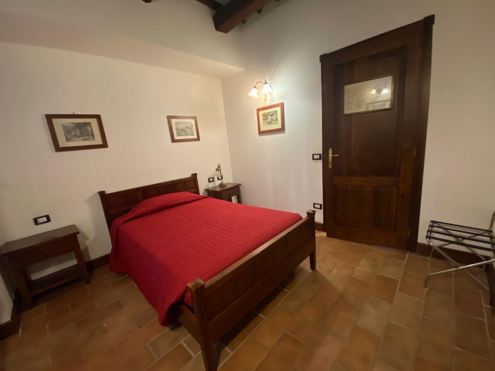 A bedroom with a wooden bed, a nightstand, a red bedspread, a door, and a wall-mounted light fixture.