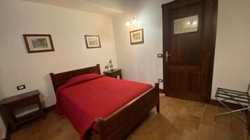 A bedroom with a wooden bed, a nightstand, a red bedspread, a door, and a wall-mounted light fixture.