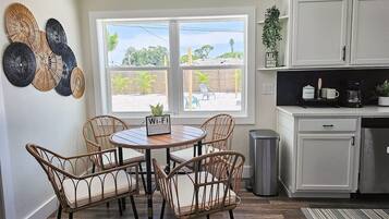 A dining area with wicker chairs and a round table, a kitchen with white cabinets, and a view of the outdoors through a large window.