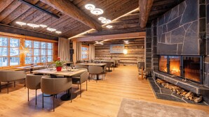 A cozy log cabin interior with a stone fireplace, wooden ceiling beams, and a dining area with tables and chairs.