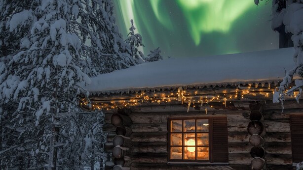 A log cabin with a lit window, surrounded by snow-covered trees and a view of the Northern Lights.