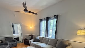 Una sala de estar con sofá, una mesa de centro, ventilador de techo, lámpara y ventana con cortinas.