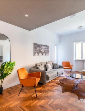 Un salon moderne avec un canapé gris, des fauteuils orange, une table basse en verre et un grand miroir.