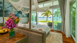 A bedroom with a bed, a bedside table with a vase of flowers, a view of the ocean, and a balcony with outdoor furniture.