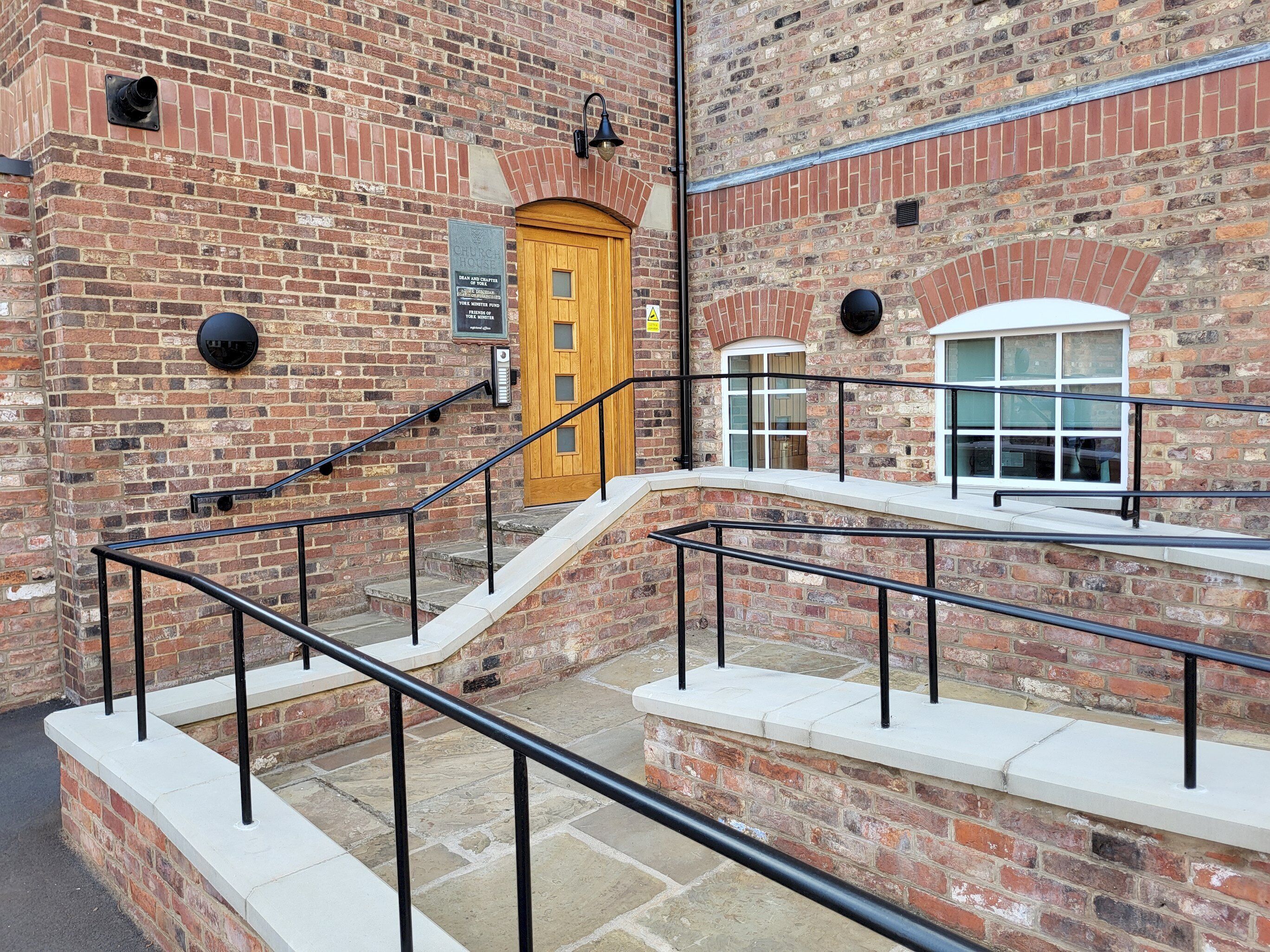 A brick building with a wooden door, a staircase with black railings, and a window with a white frame.