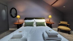A neatly made bed with white linens and green patterned pillows, two bedside lamps, a wooden chair, and a round mirror on the wall.